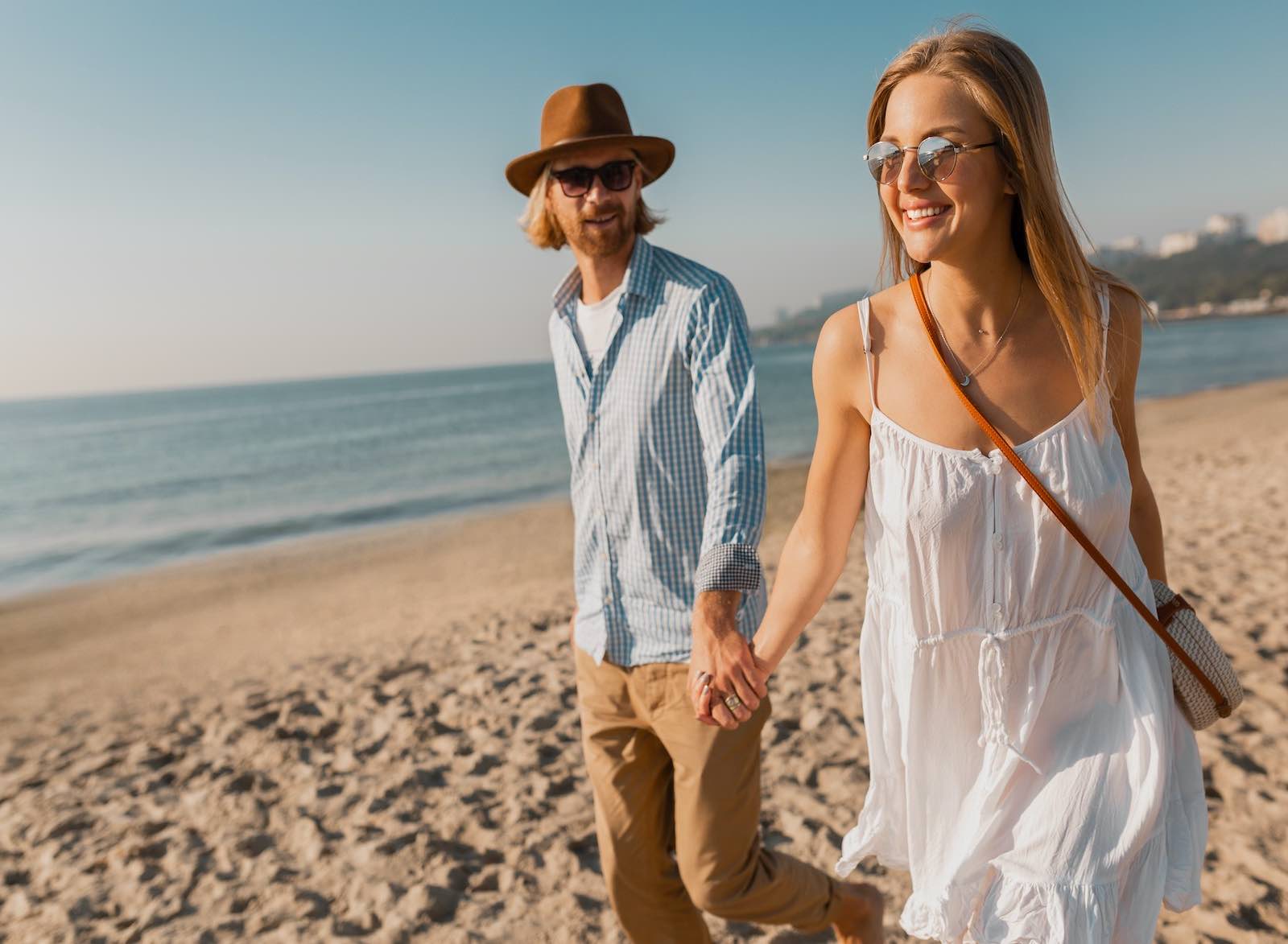 young attractive smiling happy man in hat and blond woman in white dress running together on beach on summer vacation traveling, romantic couple by the sea on sunset, stylish outfit wearing sunglasses